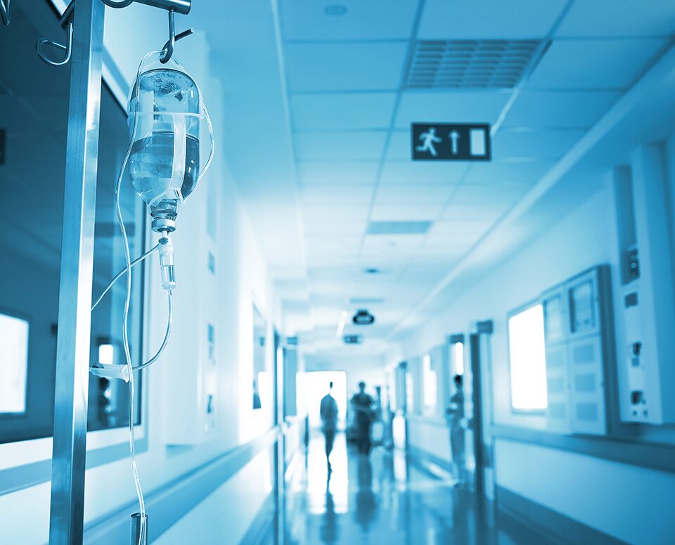 A long hospital hallway stretching into the distance, with dedicated staff members barely visible in the distance. On the left side, an IV pole stands, symbolizing medical care and treatment within the healthcare environment.