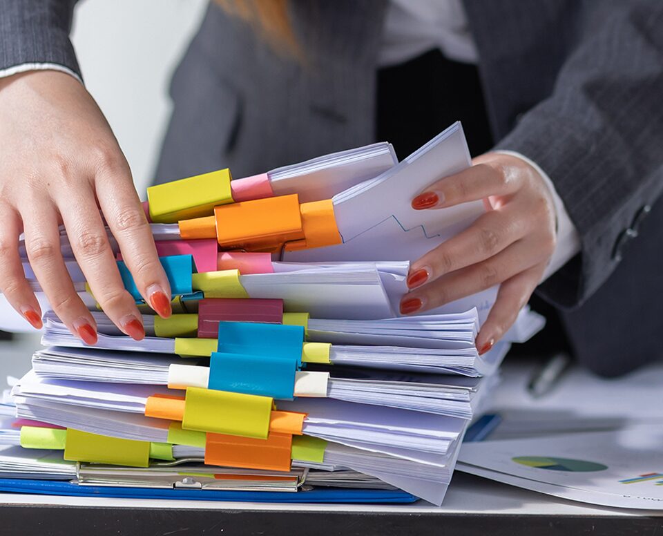 A woman meticulously sorting through a stack of papers held together by a vibrant assortment of yellow, orange, pink, blue, and white binder clips. The image showcases organizational efforts and attention to detail in managing documents and information.