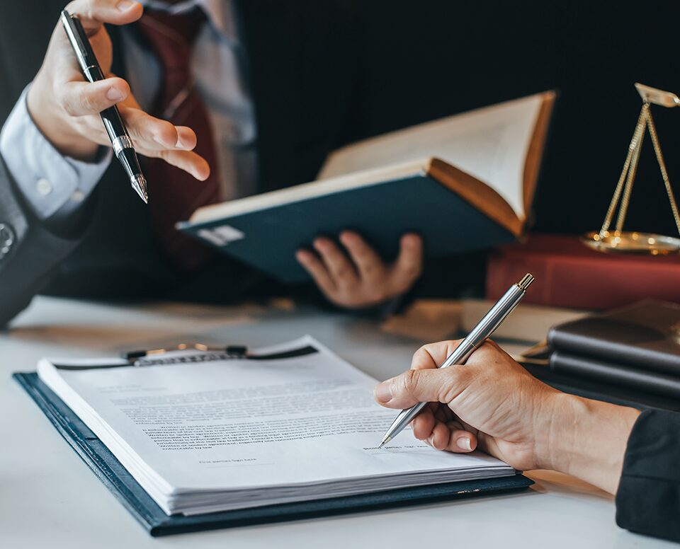 Two attorneys seated at a desk, engaged in legal activities. One attorney holds a book, showcasing legal knowledge, while the other attorney is signing a document, representing the act of finalizing legal agreements and contracts.